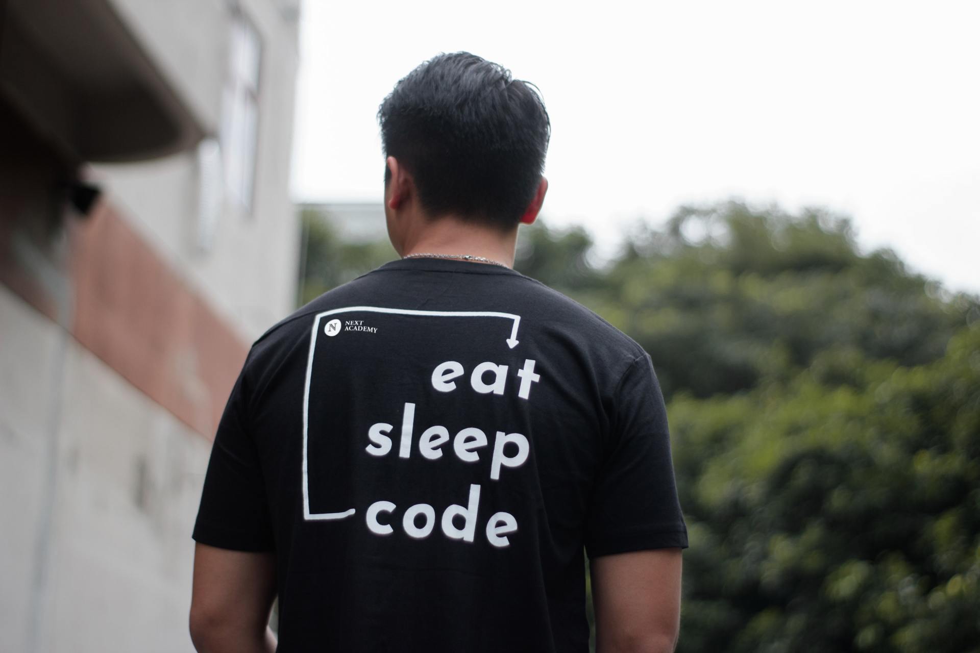 A man wearing a black t-shirt that says `` eat sleep code '' is standing in front of a building.