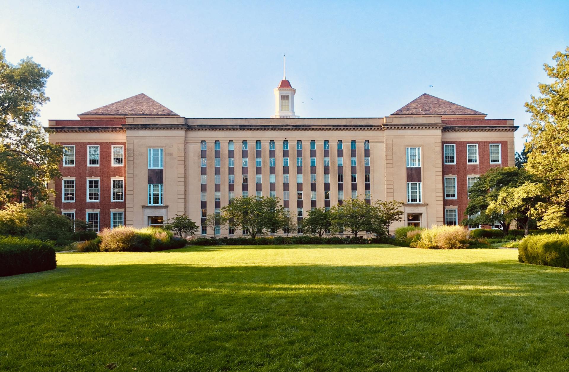 A large brick building with a clock tower in the middle of a lush green field.
