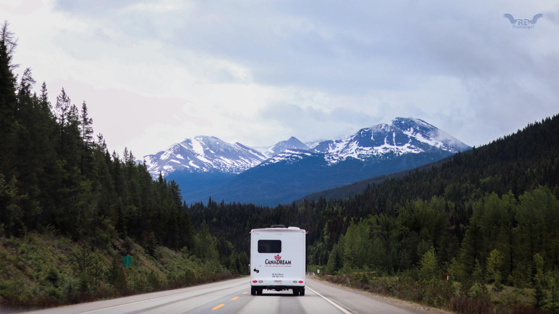 A camper van drives down a highway with snow-capped mountains in the background.