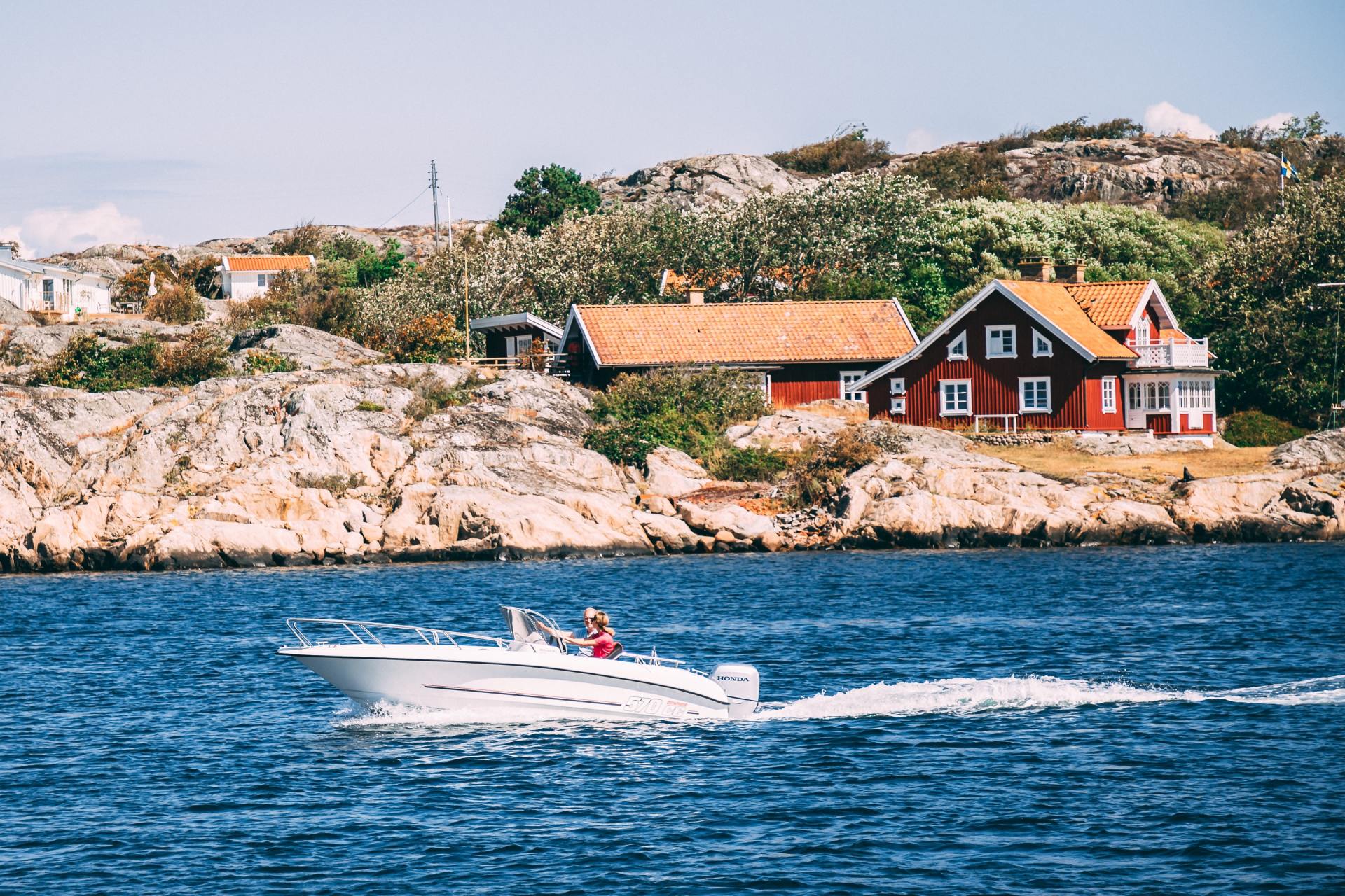 White speedboat with two people on blue water near rocky shore and red houses