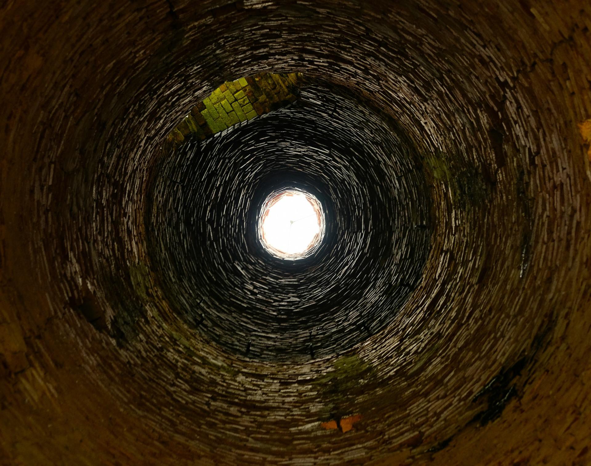 Looking up from inside a dark, cylindrical well at bright daylight; brick walls.
