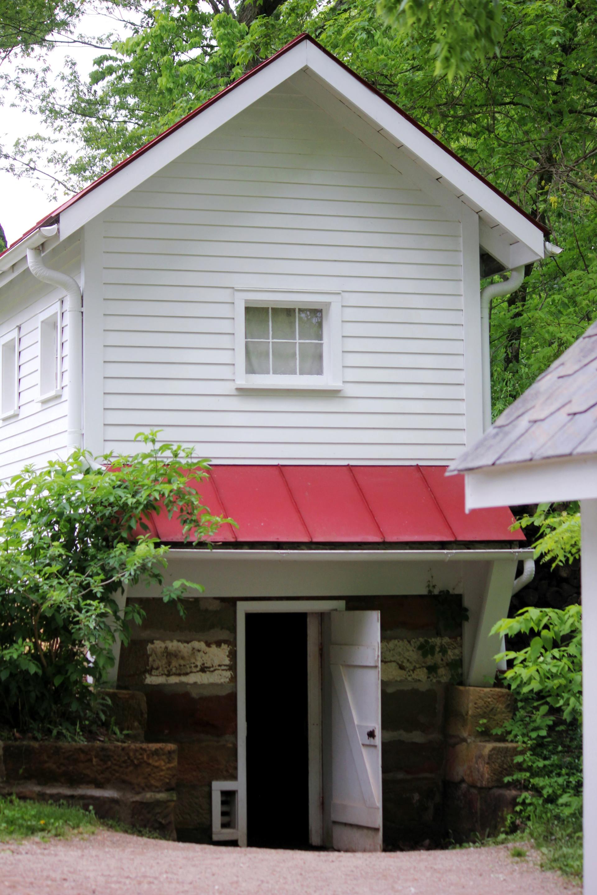 A white wooden building with a red metal awning over a stone-walled entrance, nestled among lush green trees.
