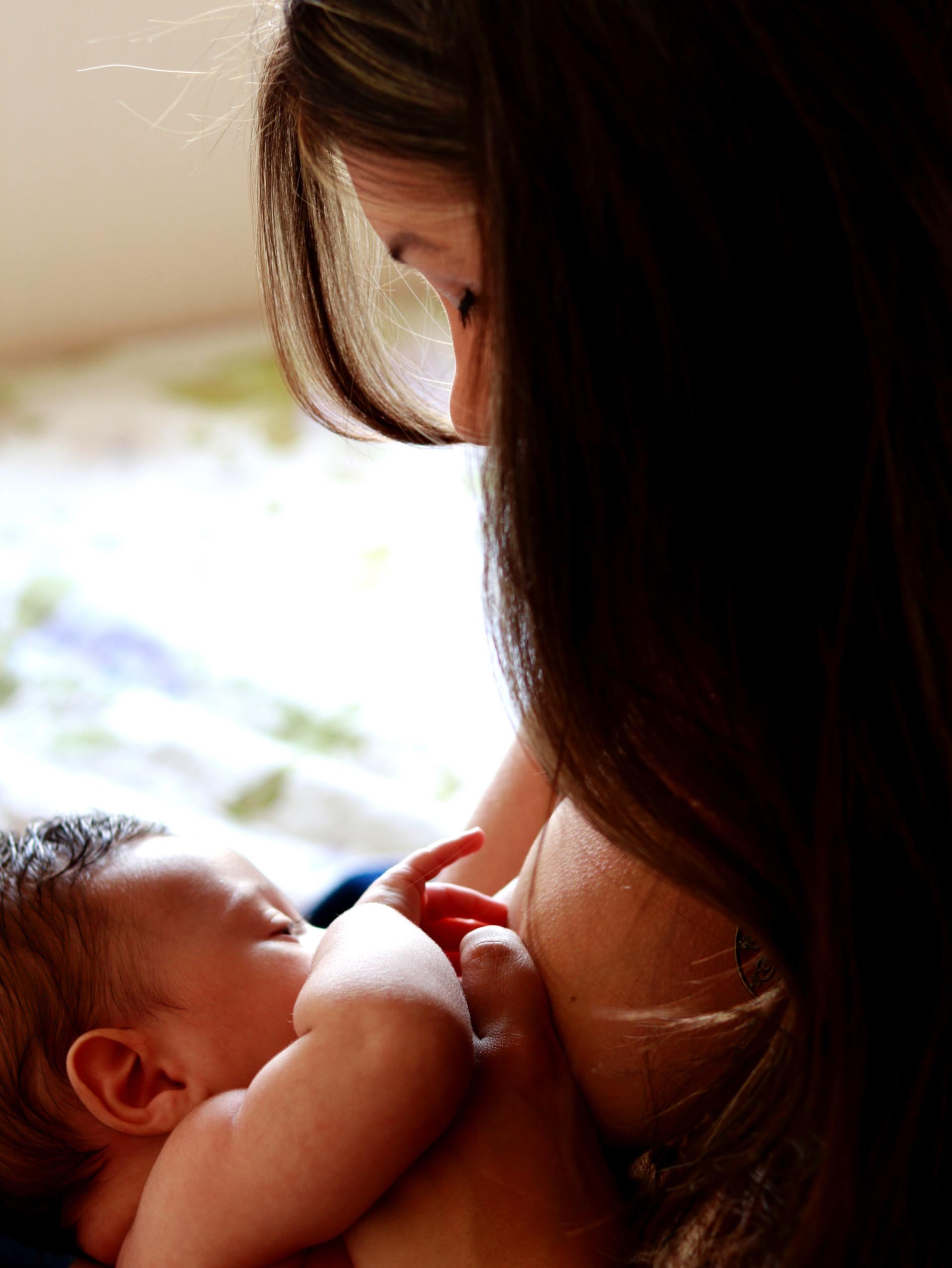 A woman with long dark hair breastfeeds a baby indoors. The baby nurses, and the woman looks down lovingly.