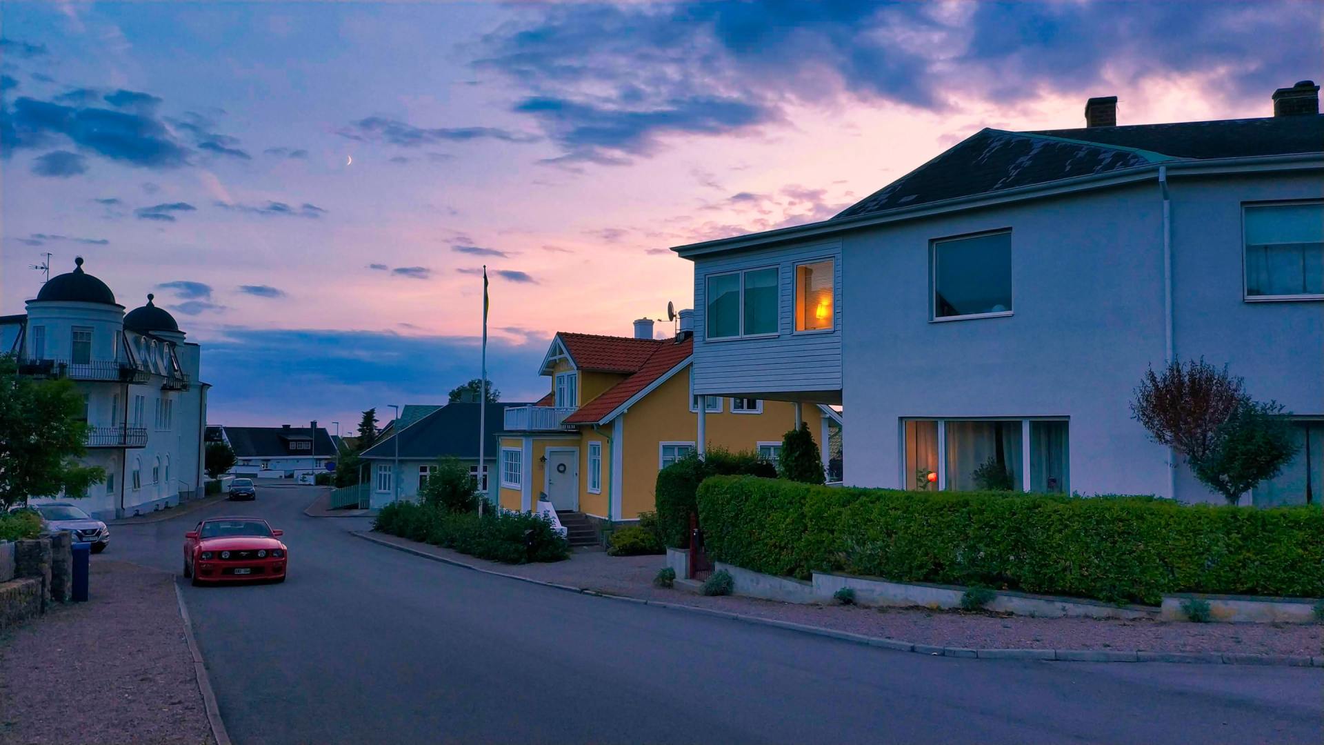 A red car is driving down a street next to a row of houses at sunset.
