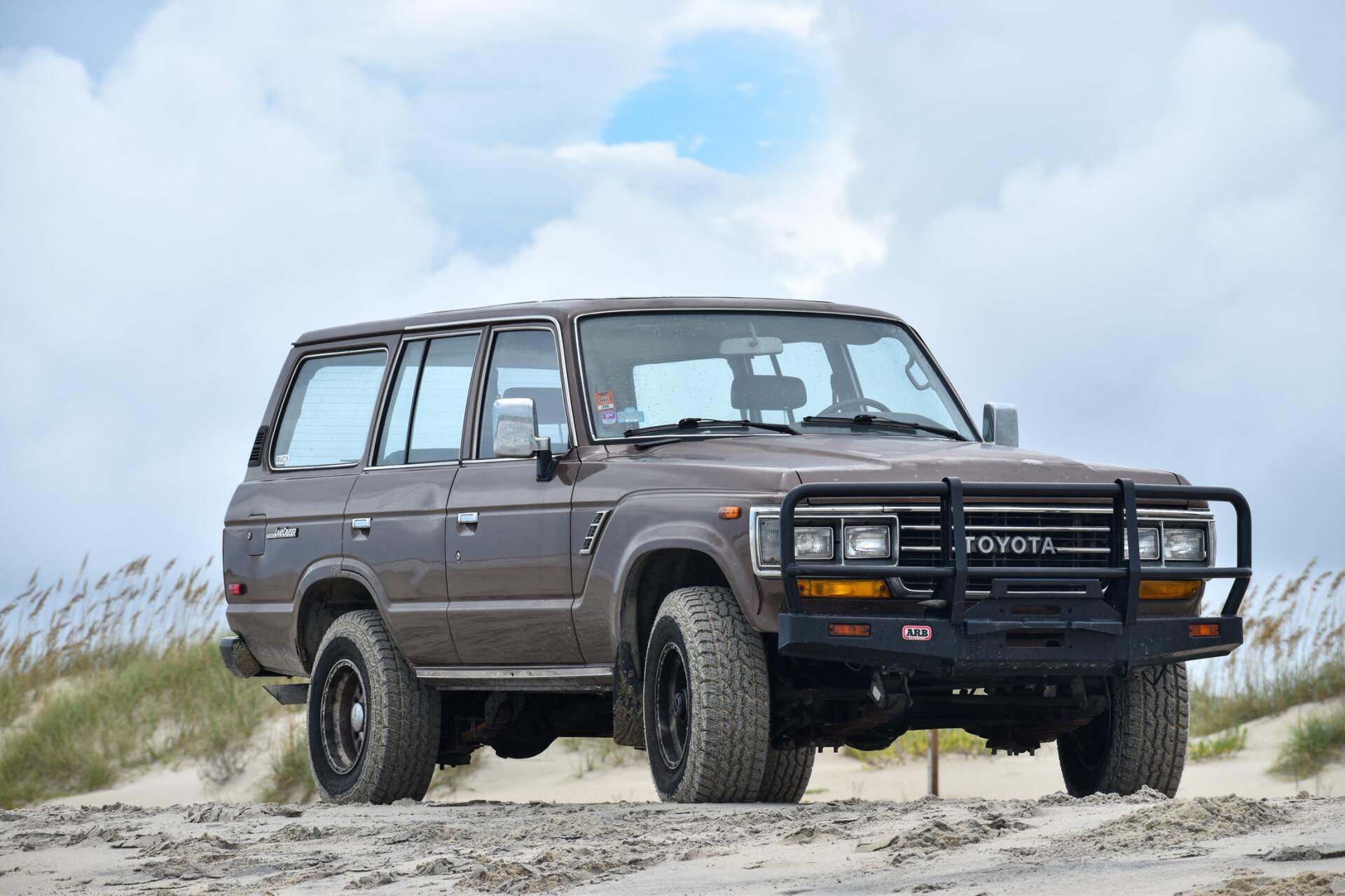 Brown Toyota Land Cruiser SUV parked on a sandy beach under a cloudy sky.