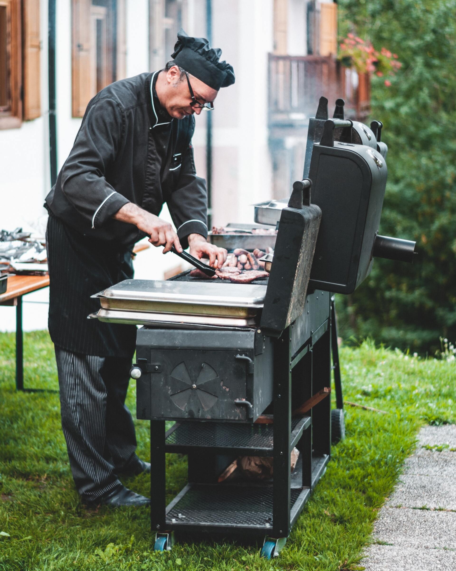 Chef grilling meat outdoors at a lodge, wearing black chef attire and glasses, using tongs.