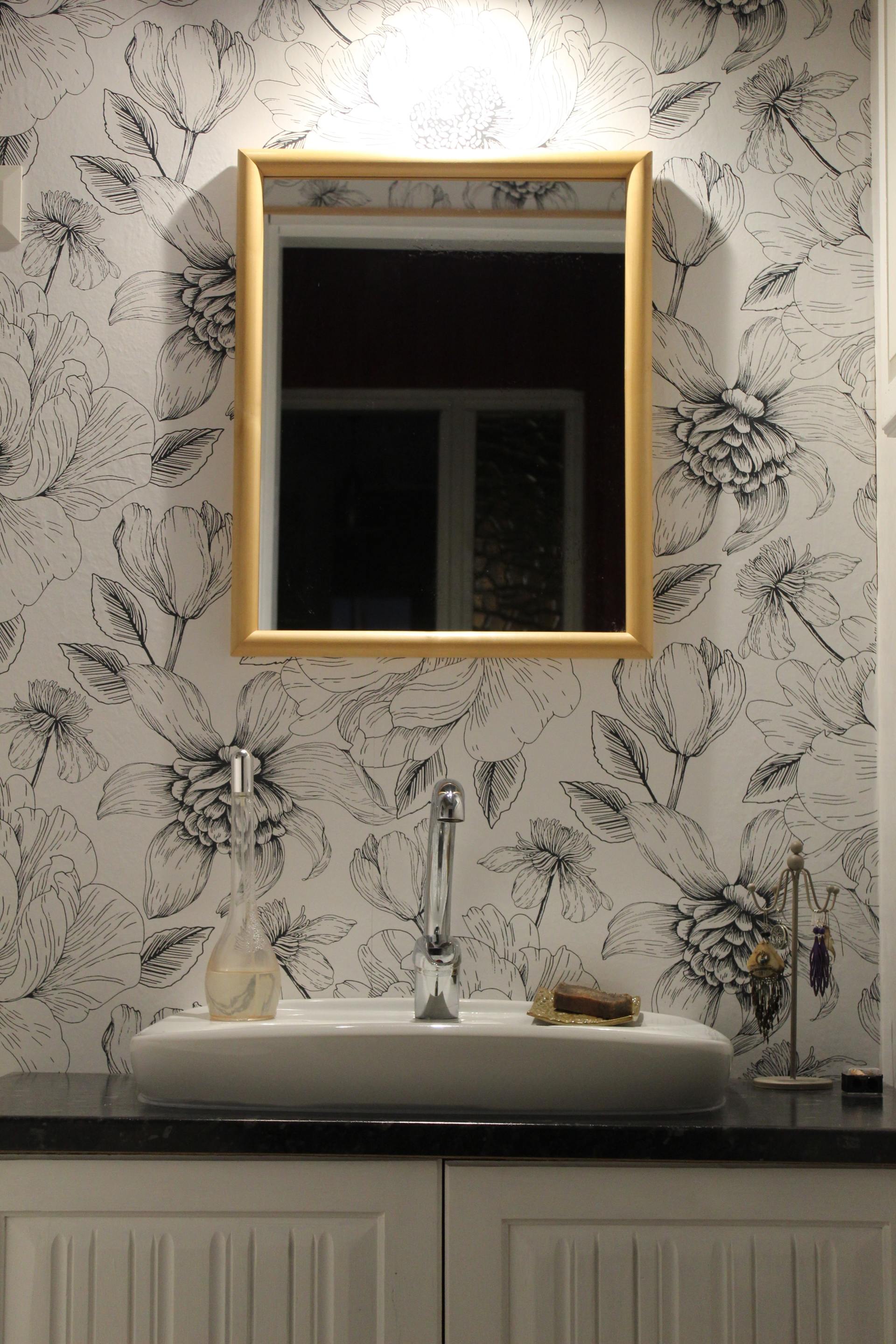 Bathroom with floral wallpaper, gold-framed mirror over a white sink and vanity.
