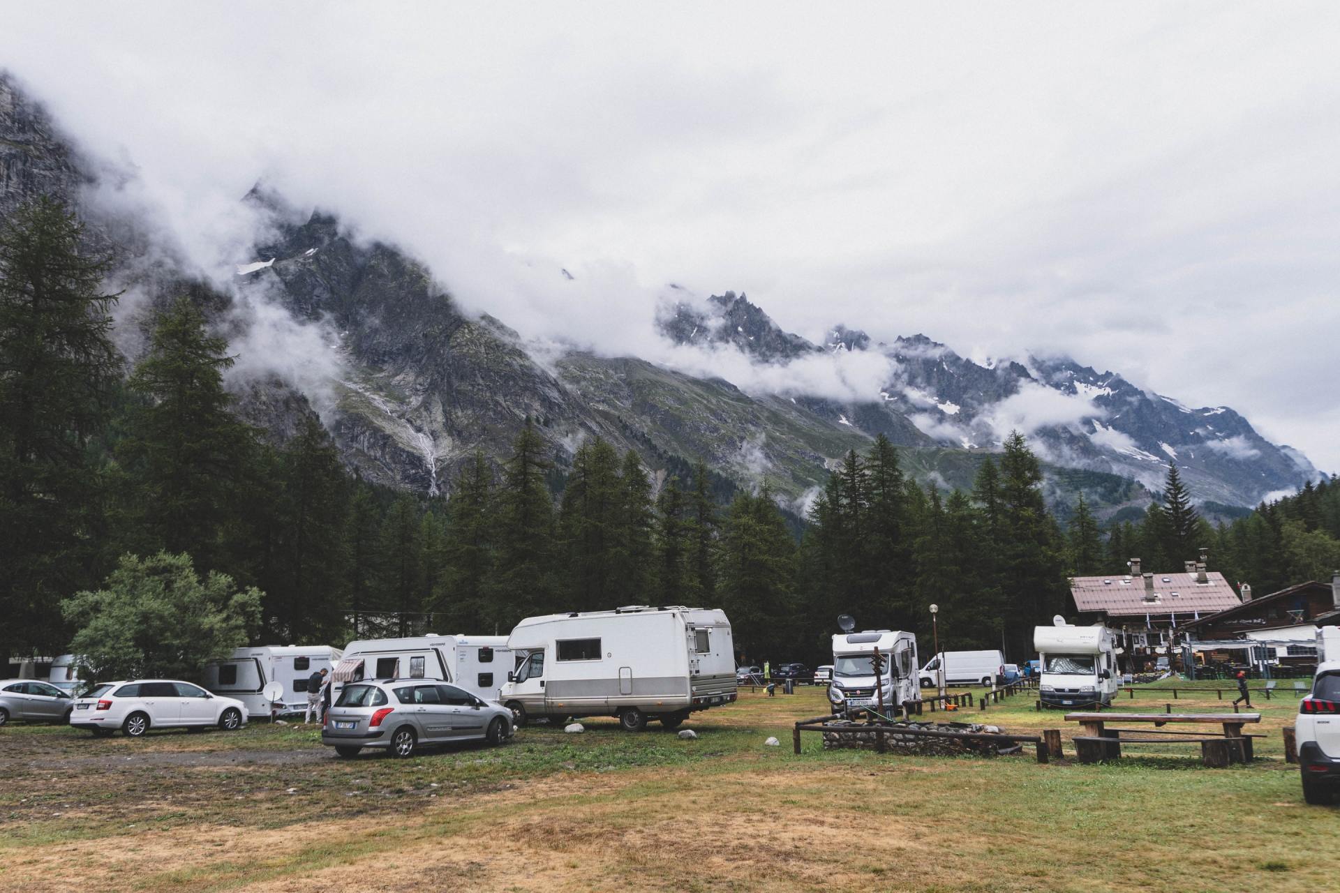 Campground with RVs and cars nestled among tall trees, set against misty mountains.