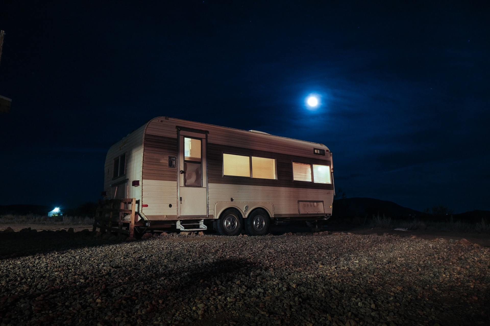 Vintage camper trailer illuminated at night under a bright moon, surrounded by gravel.