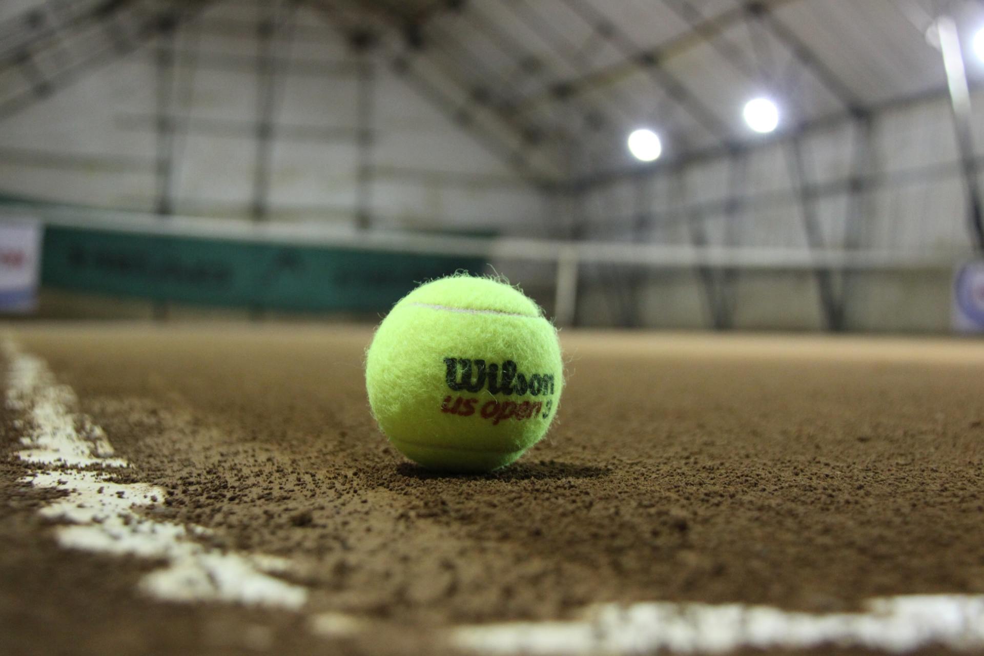 A wilson tennis ball is sitting on the edge of a tennis court. of an indoor sports hall needing heating