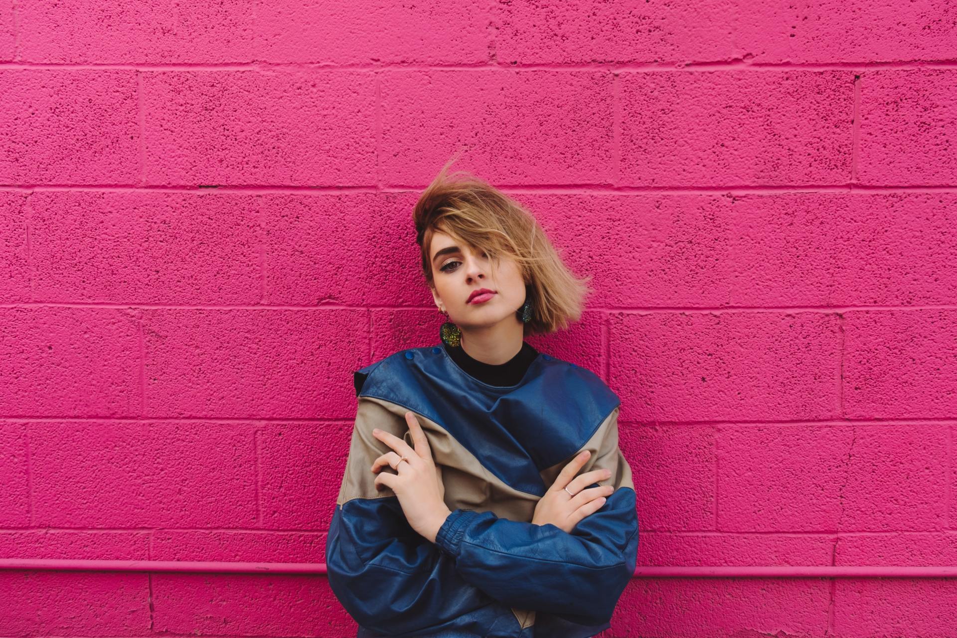 A woman in a blue jacket is standing in front of a pink brick wall.
