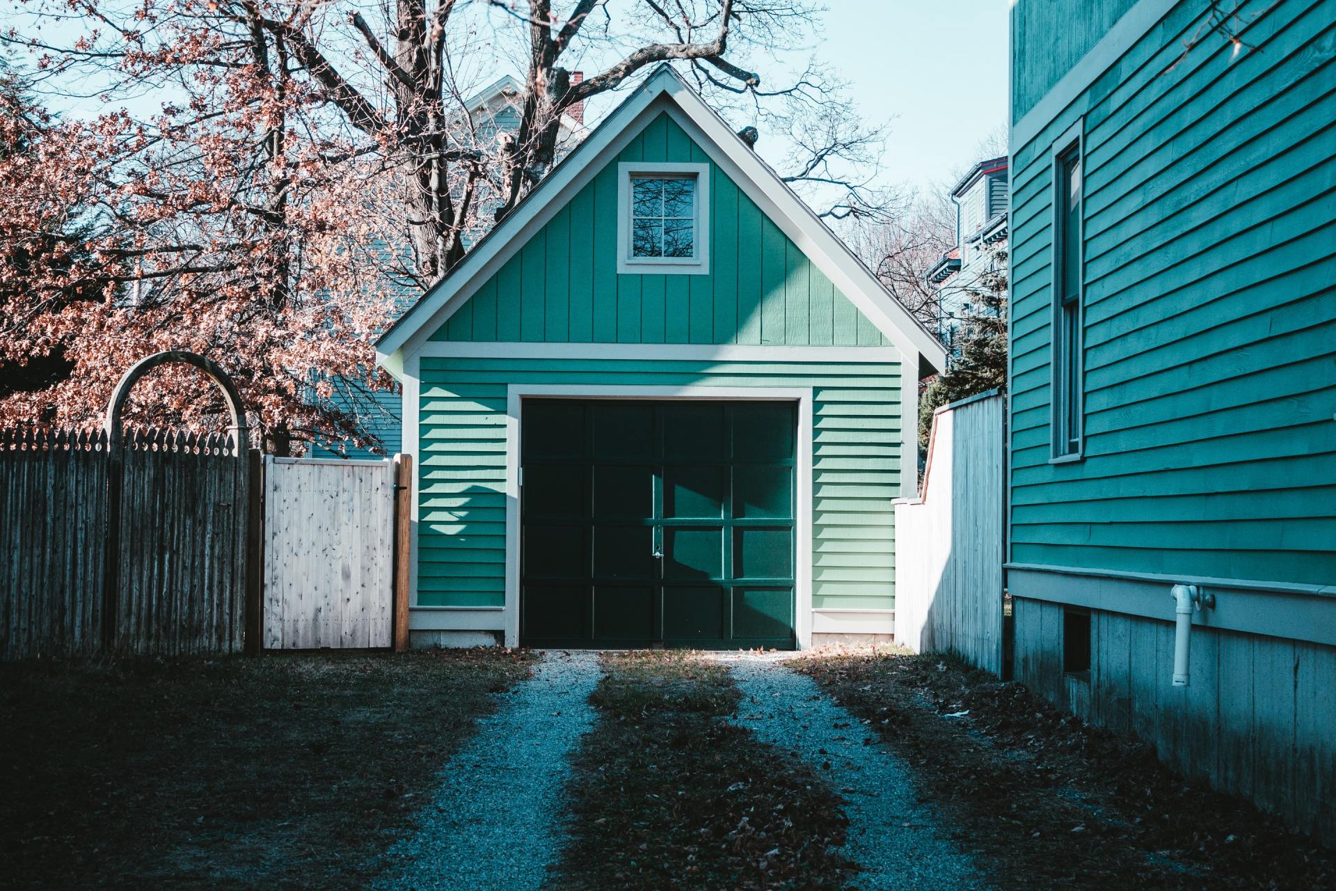 A green garage with a driveway leading to it is next to a blue house.