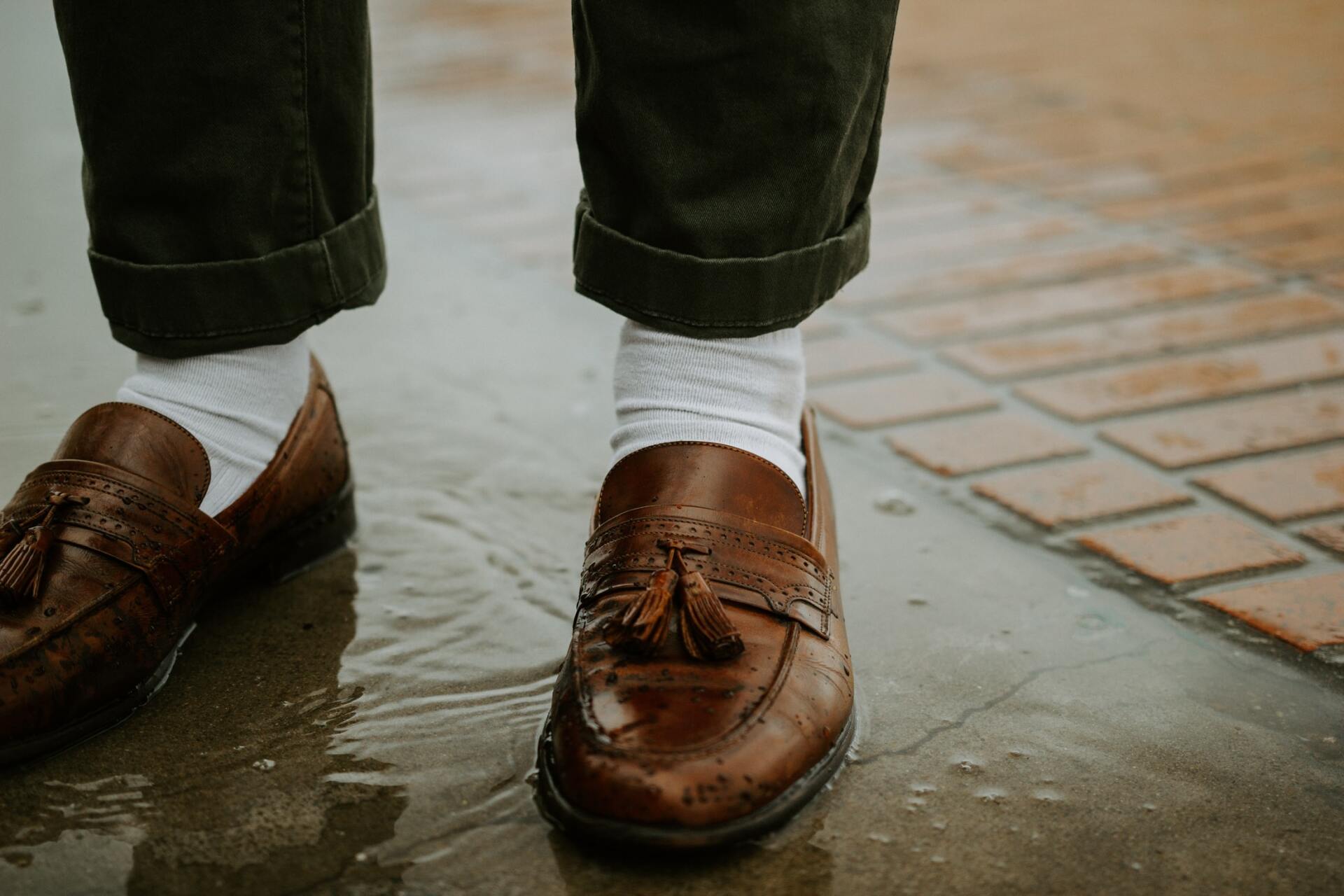 A person wearing brown loafers and white socks is standing in a puddle of water.