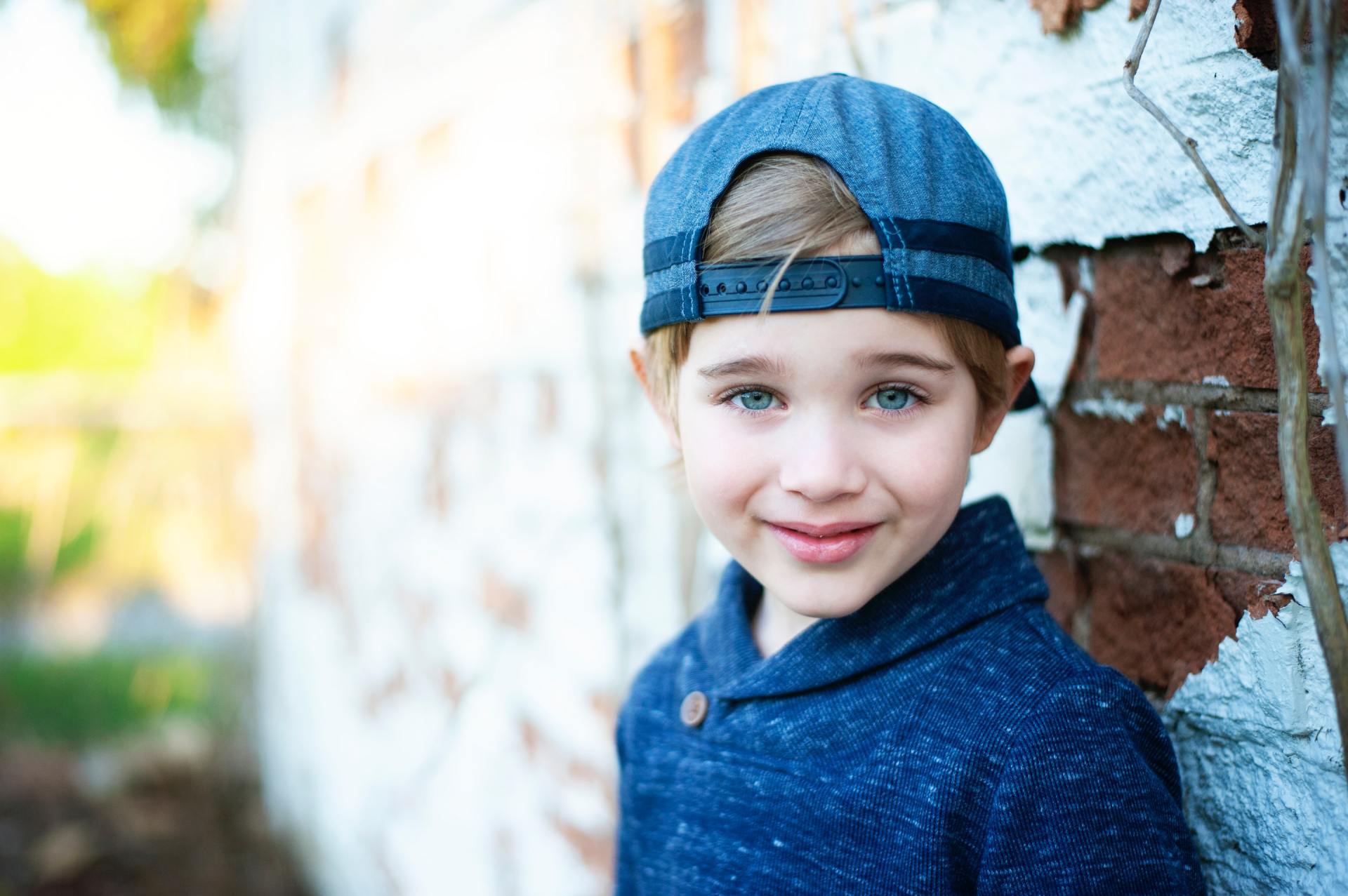 A young boy wearing a baseball cap and a blue sweater is leaning against a brick wall.