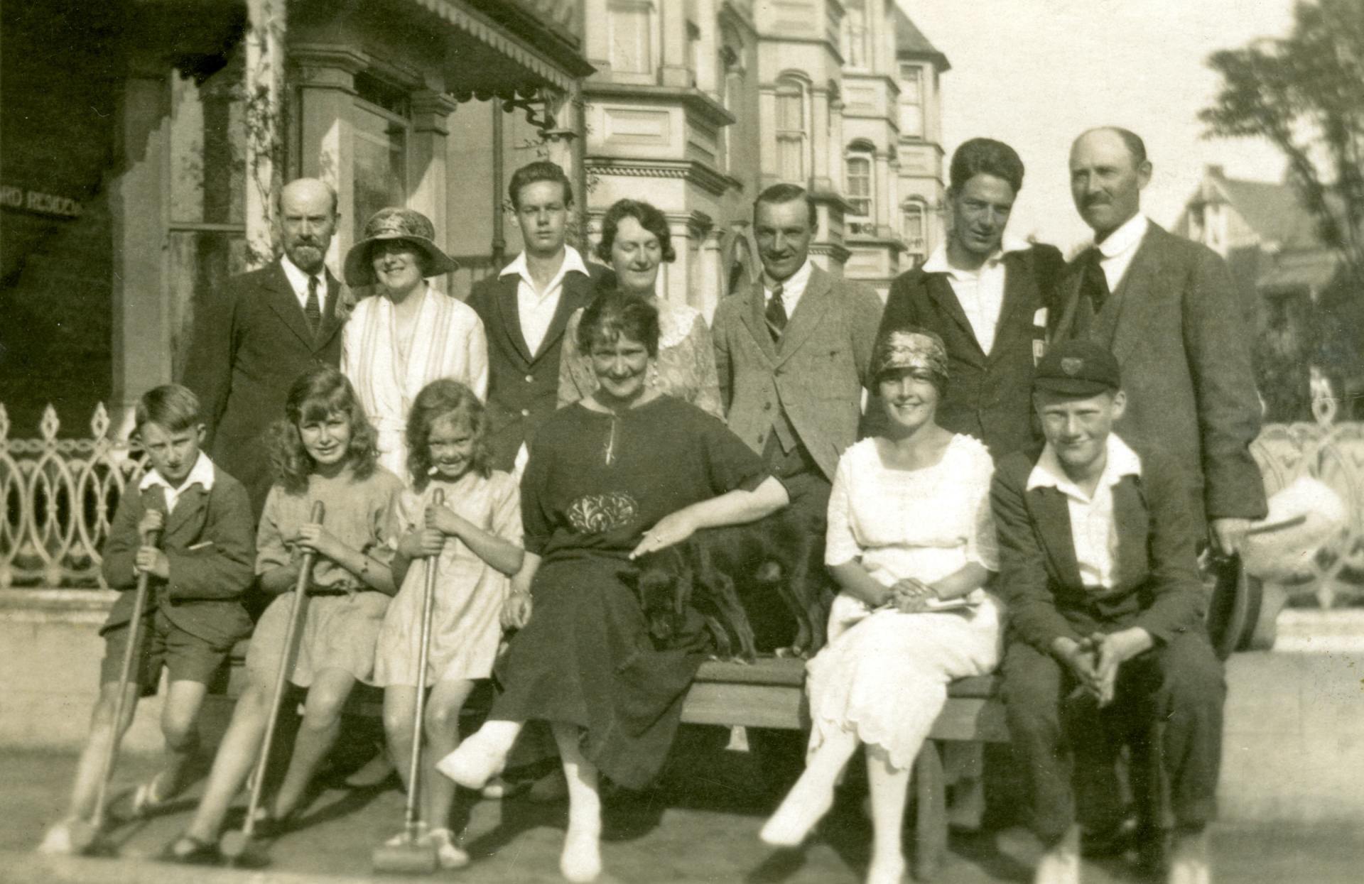 A group of thirteen people standing and sitting together outside a building, dressed in formal vintage clothing.