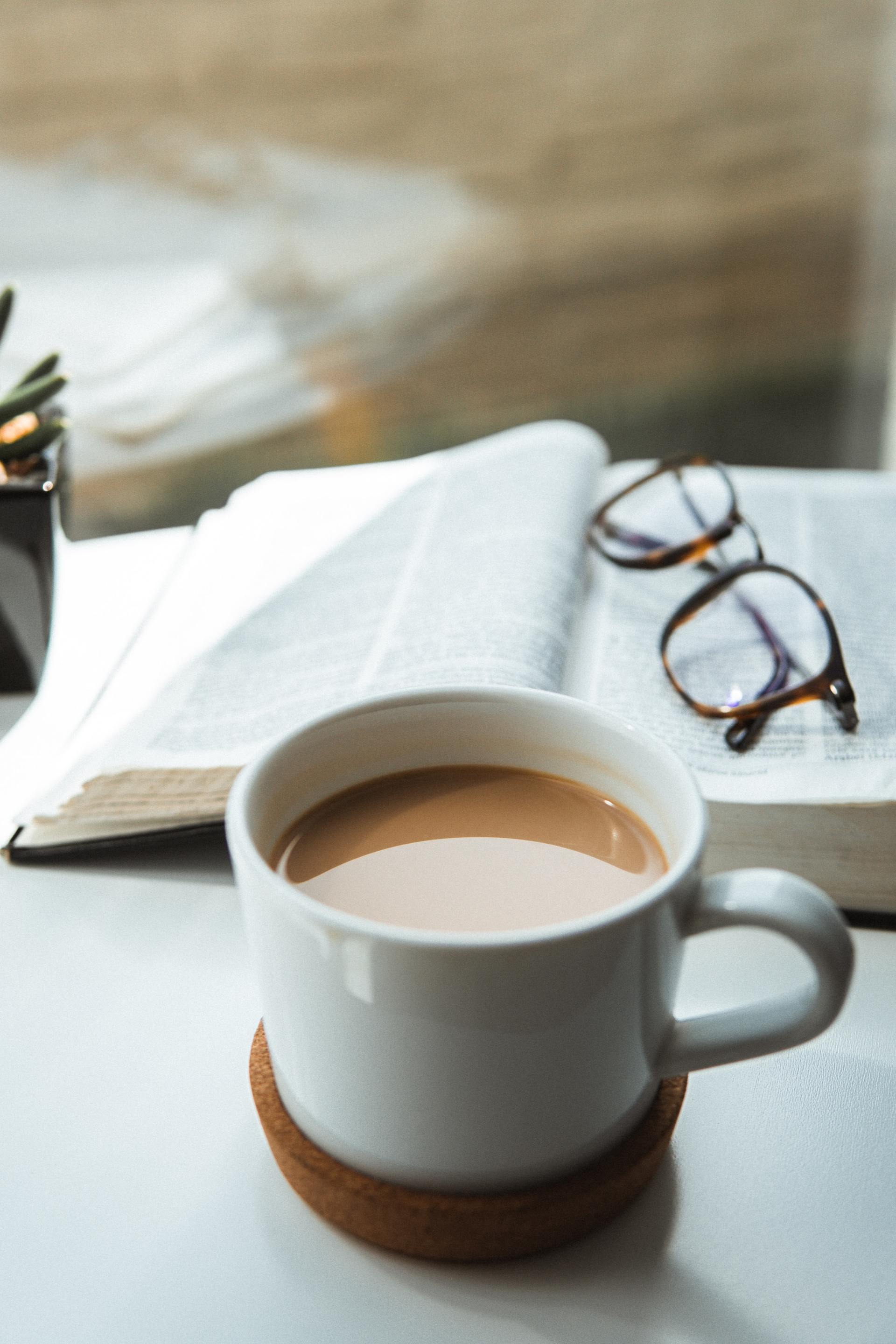 A cup of coffee is sitting on a table next to a book and glasses.