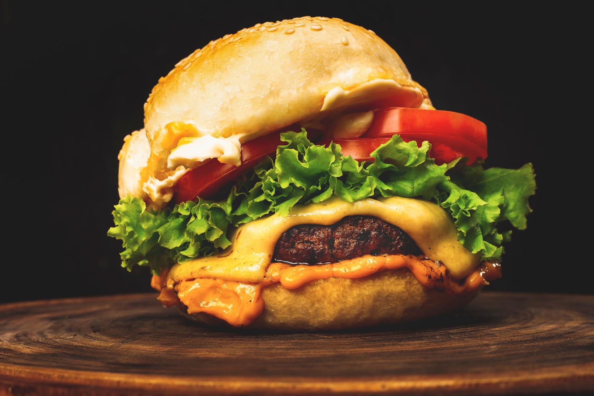 Burger with melted cheese, lettuce, tomato, and sauce on a sesame seed bun, on a wooden surface, black background.