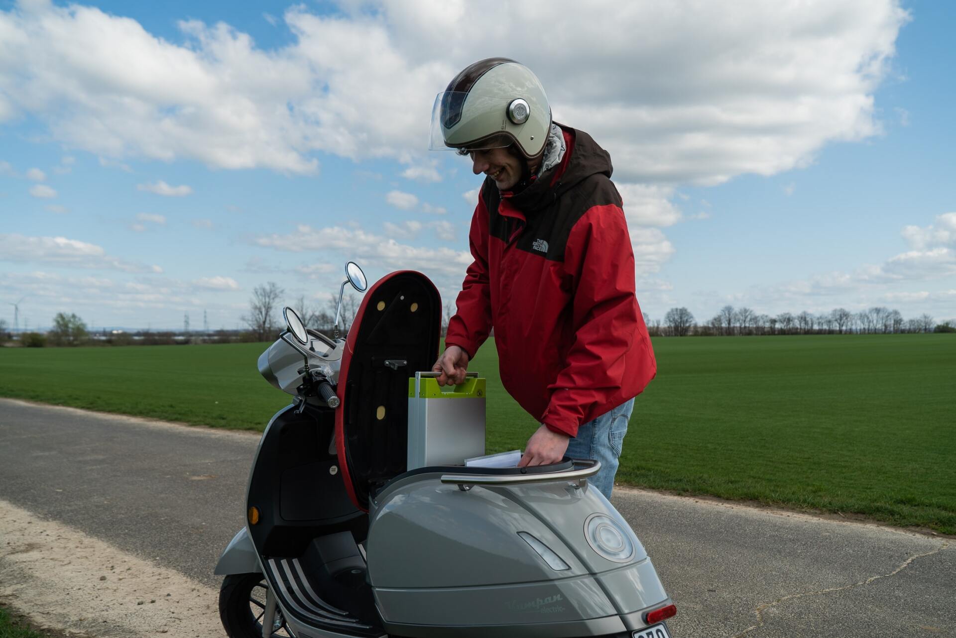 Person in red jacket and helmet reaching into storage on a gray scooter, next to a field.