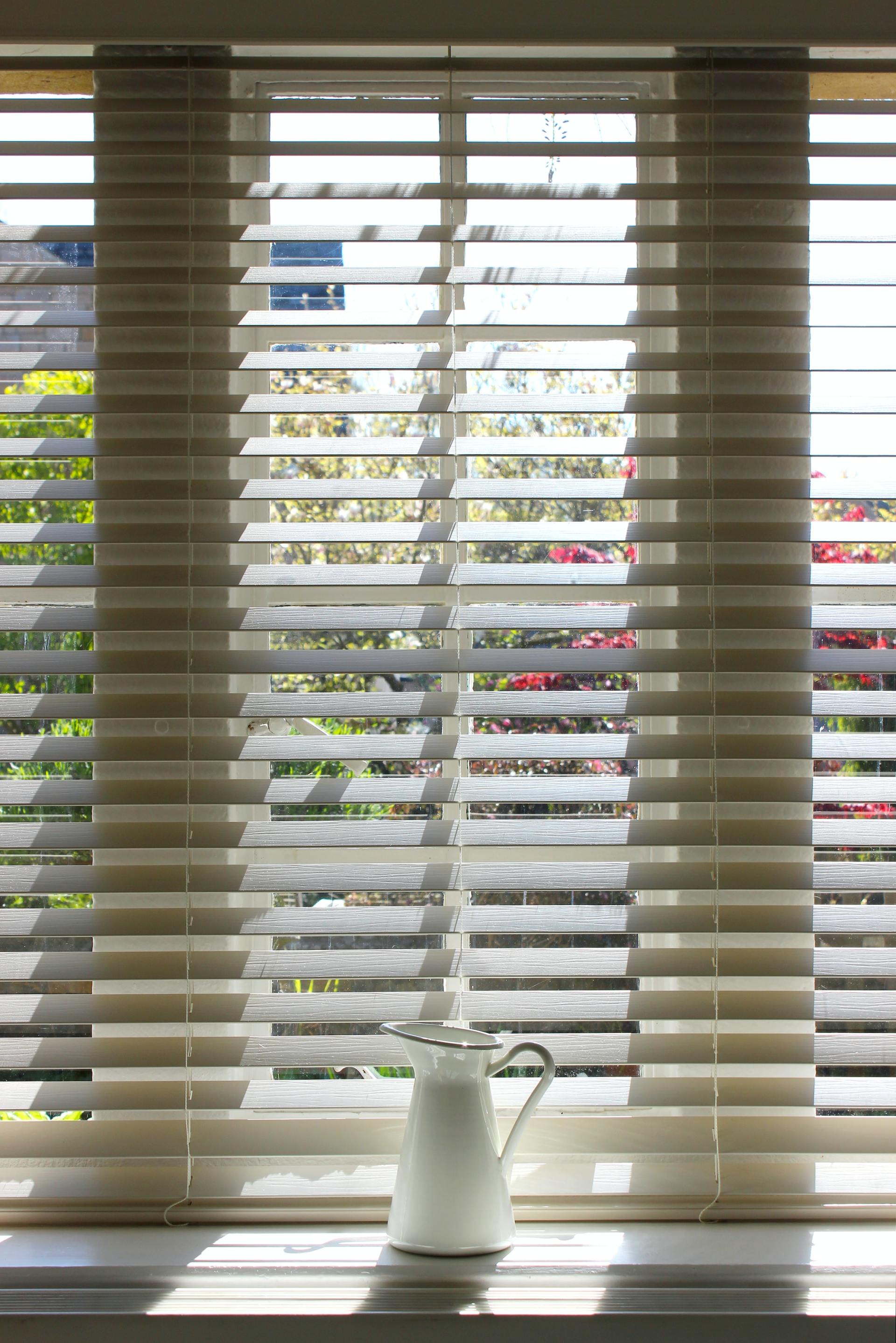 A white pitcher is sitting on a window sill next to a window with blinds.