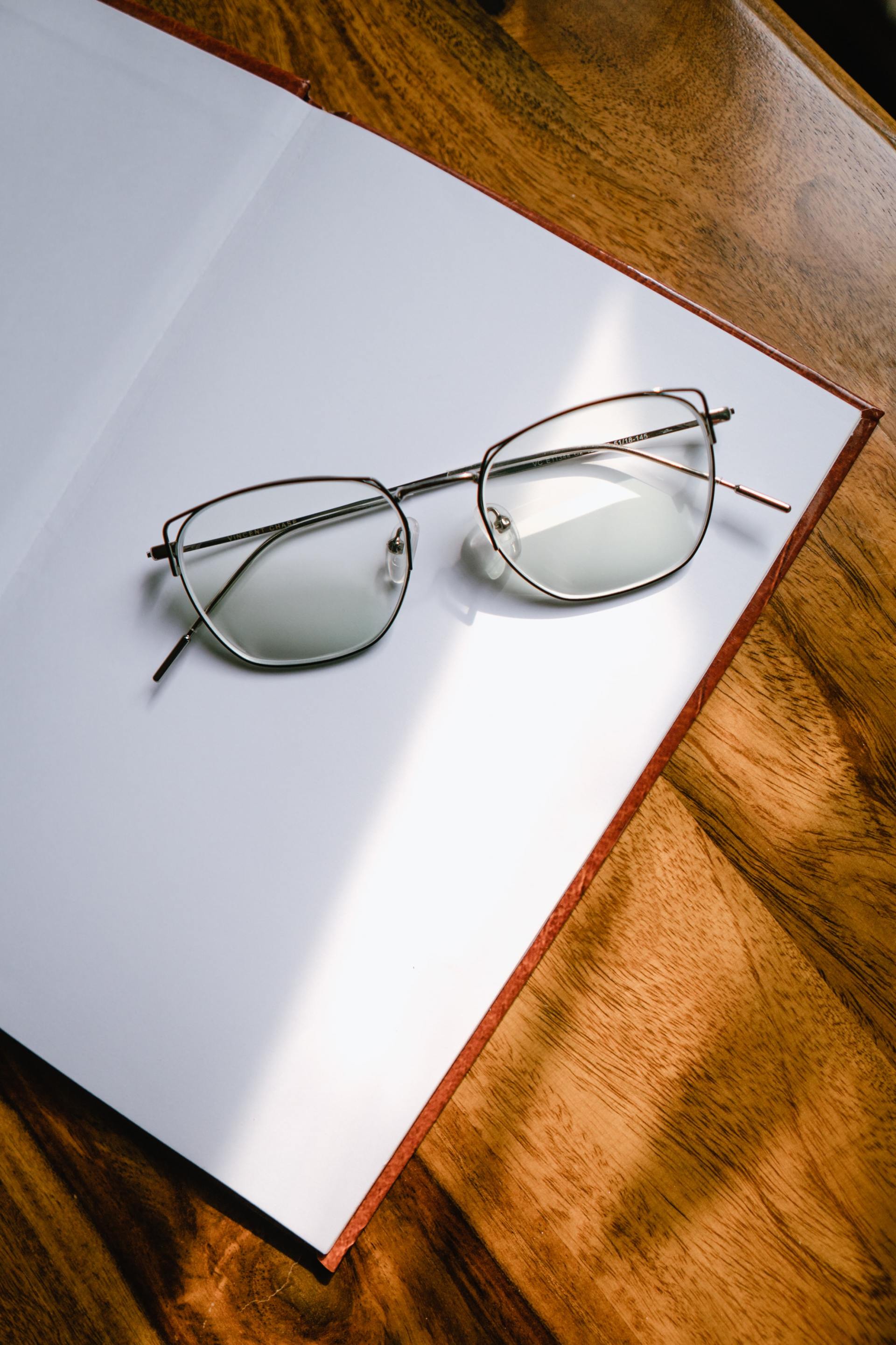 Eyeglasses resting on an open notebook on a wooden table, bathed in sunlight.