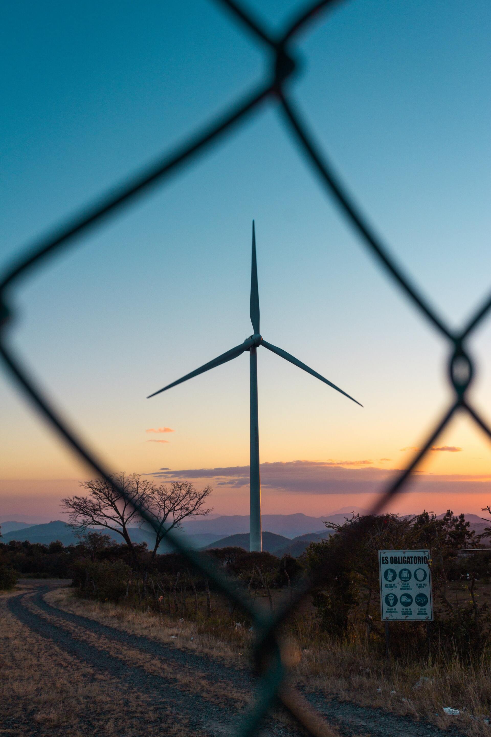 A wind turbine is behind a chain link fence at sunset.