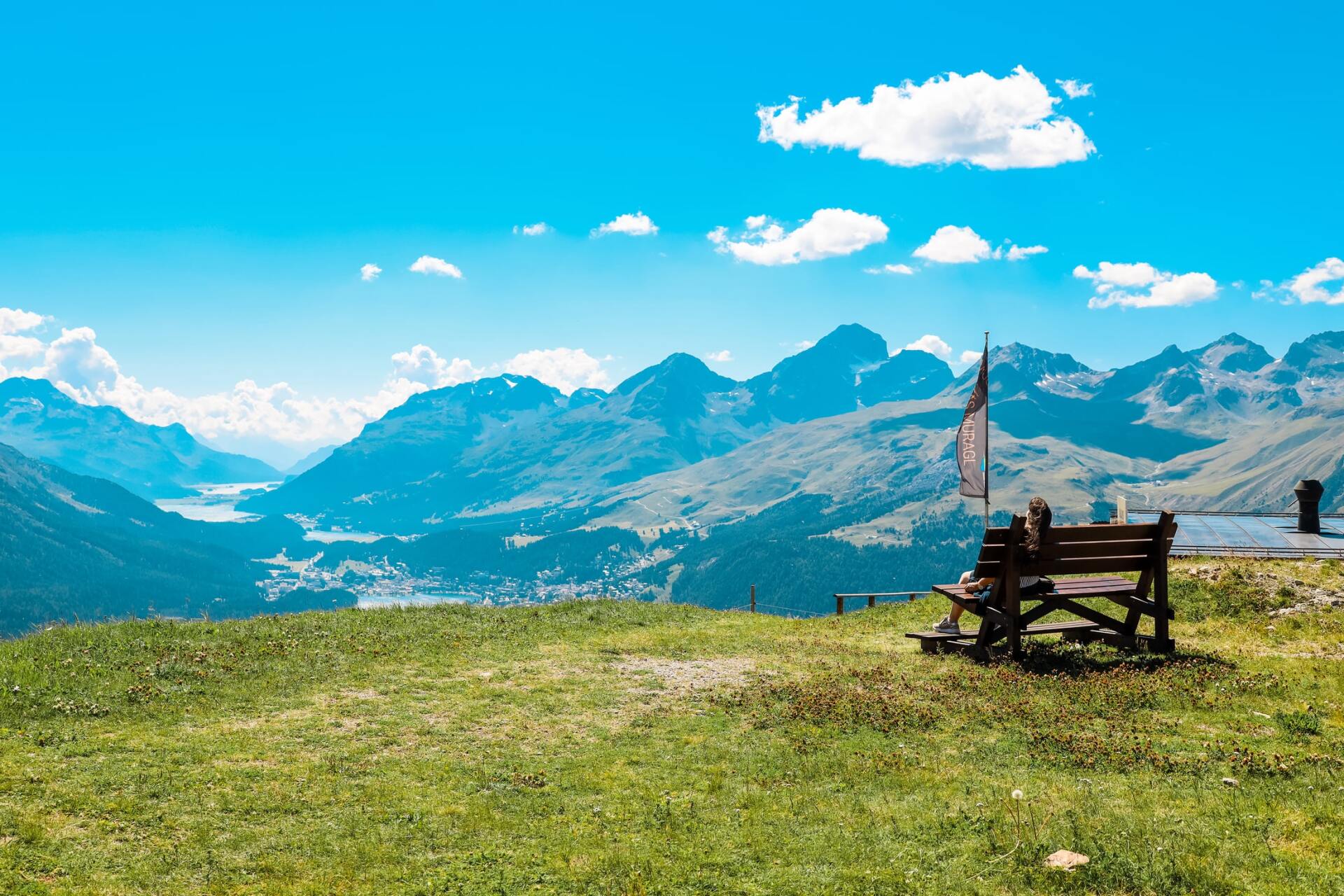 Eine Holzbank steht auf einem grasbewachsenen Hügel mit Aussicht auf eine Bergkette.