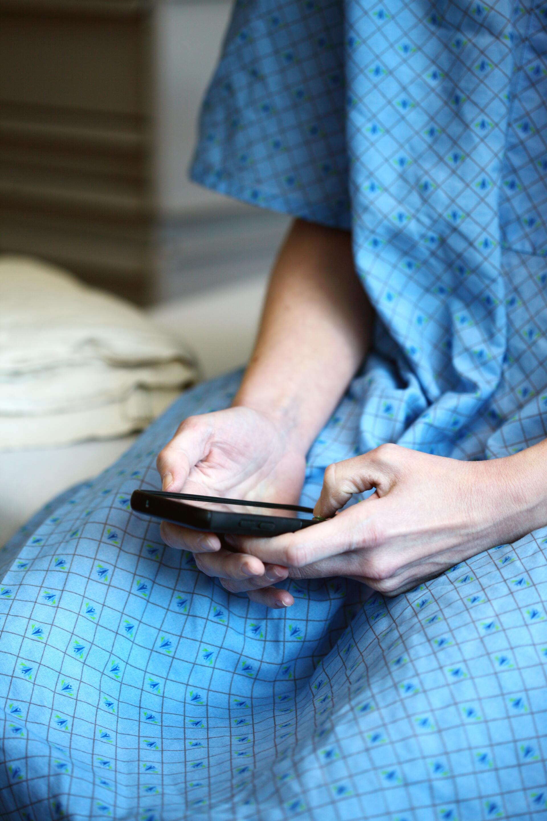 Person in a blue hospital gown holding and looking at a smartphone, seated on a bed.