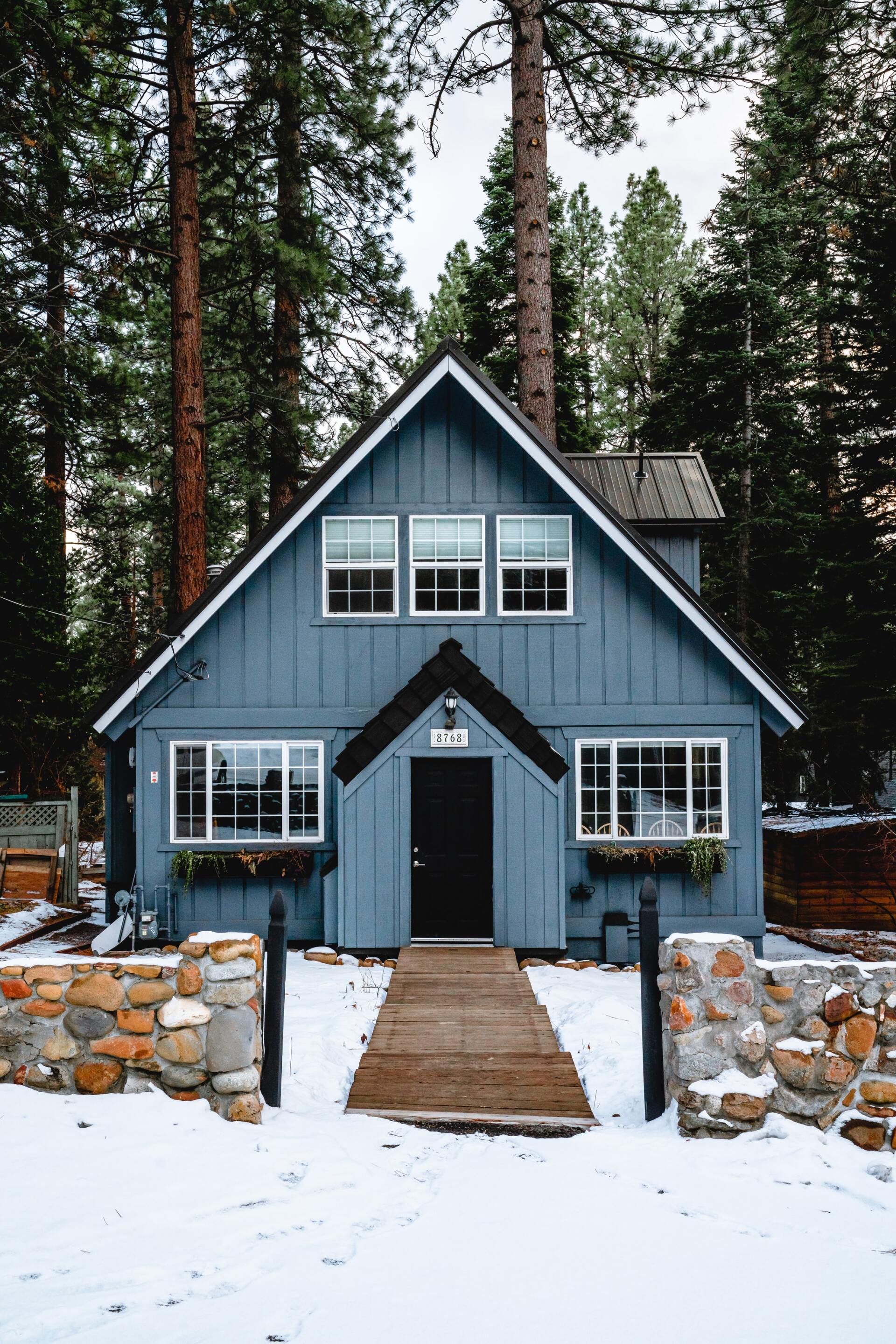 Blue cottage with a black door, surrounded by snow and tall pine trees.