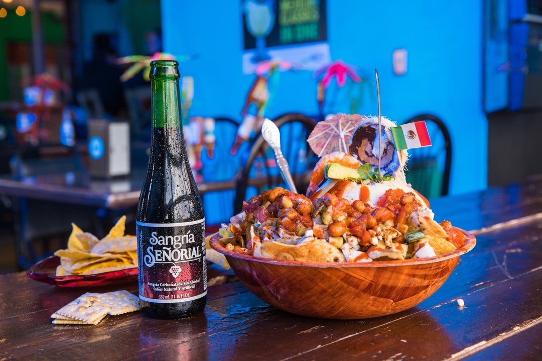 A large bowl of nachos with a beer bottle and chips on a bar. Mexican flag decorates the nachos.