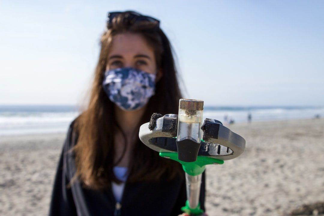 A woman wearing a mask is holding a green object on the beach.