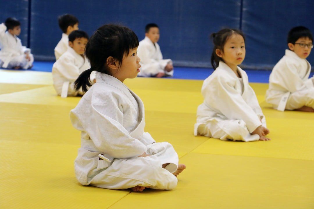 Een groep jonge kinderen in karate-uniformen zit op een gele mat.